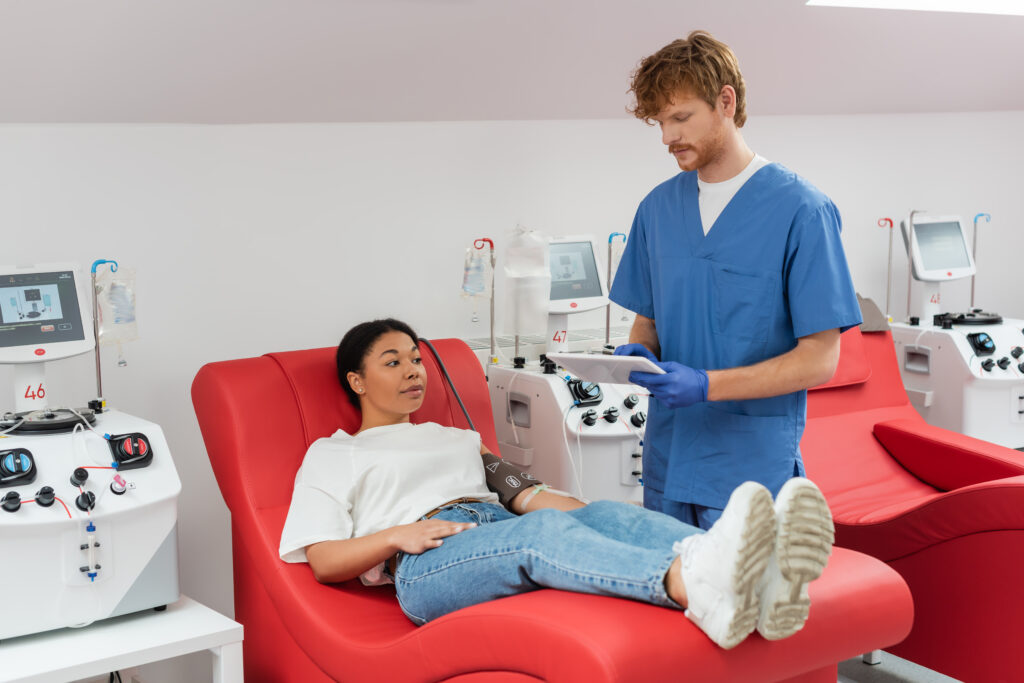 redhead doctor in blue uniform and latex gloves using digital tablet near transfusion machine and multiracial woman with pressure cuff sitting on medical chair in clinic
